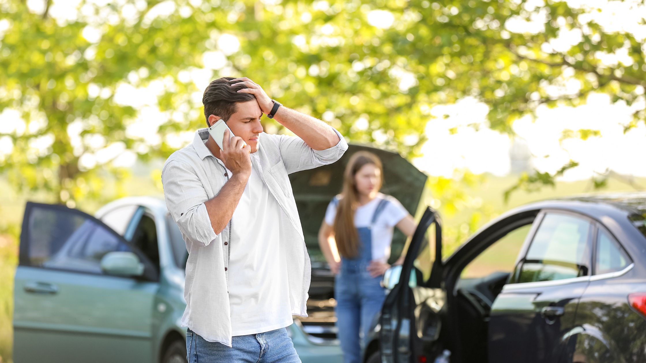 A distressed man on the phone after a vehicle collision, potentially discussing options with a Balcones Heights car accident lawyer, with an open car hood and a woman in the background.